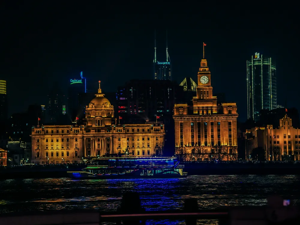 Un barco de crucero iluminado en azul sobre el río frente a los edificios históricos del Bund bañados en oro y su torre del reloj, de noche