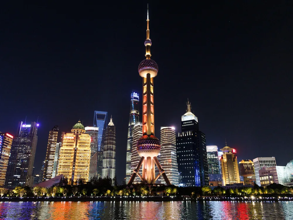 La Torre de la Perla Oriental iluminada en dorado y naranja sobre el paseo marítimo de Pudong, vista desde el río, con otras torres a su alrededor